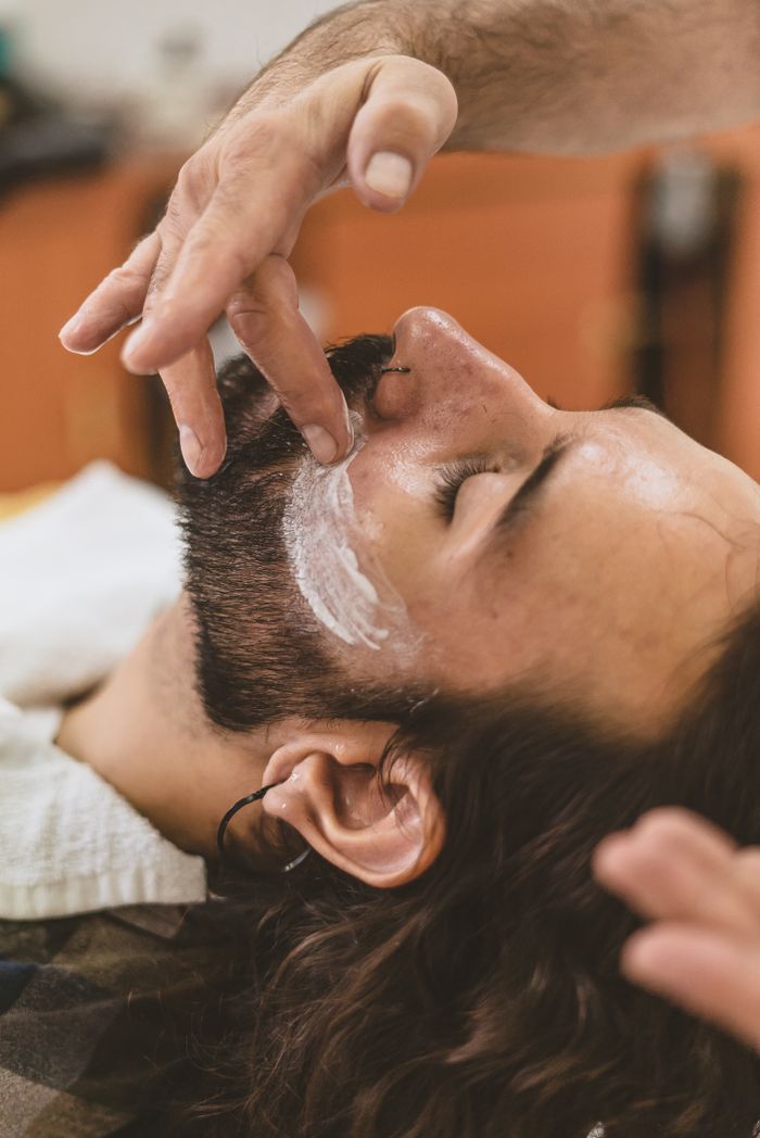 A man with long hair receives a beard trim and shaving cream application at a barber shop.